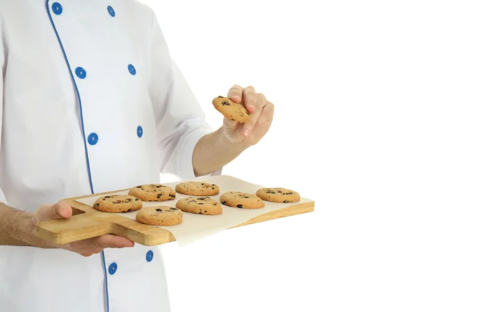 A home baker looking puzzled while examining a tray of flat chocolate chip cookies, with a kitchen background.