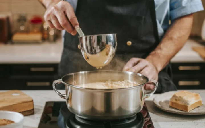 A chef whisking a roux in a pot on the stove, with cornstarch and cheese on the countertop, demonstrating thickening techniques for soup.