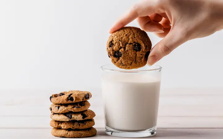 mini chocolate chip cookies scattered around a small plate, with a glass of milk in the background.