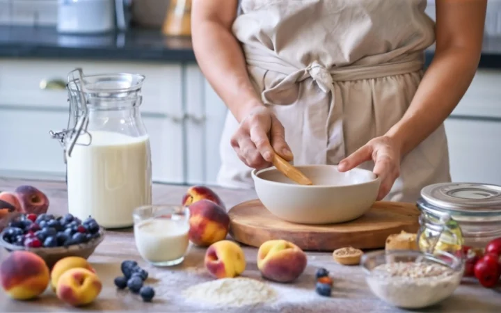 A kitchen counter with various high-quality baking ingredients, including fresh fruits like peaches and berries, homemade dessert mix, and organic butter. A baker's hand is seen selecting ingredients.