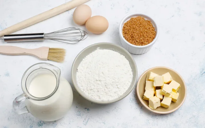  Close-up of various baking ingredients like flour, sugar, and butter arranged neatly on a kitchen counter, with a recipe book open in the background.