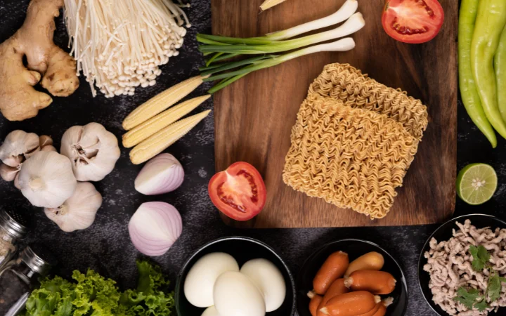 noodles on a wooden cutting board with tomato, lime, spring onion, chili and baby corn