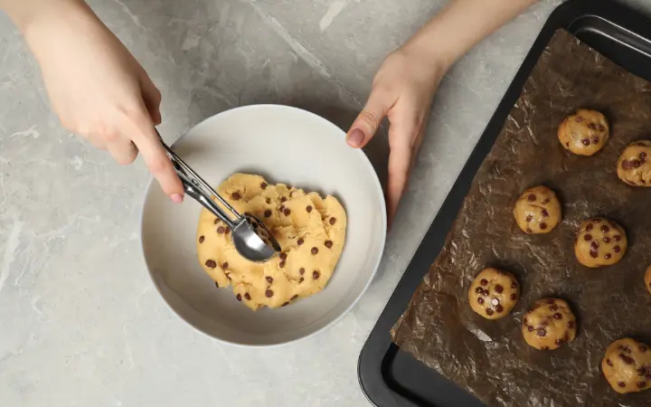 Hands preparing mini chocolate chip cookie dough in a mixing bowl, with a wooden spoon and ingredients visible.