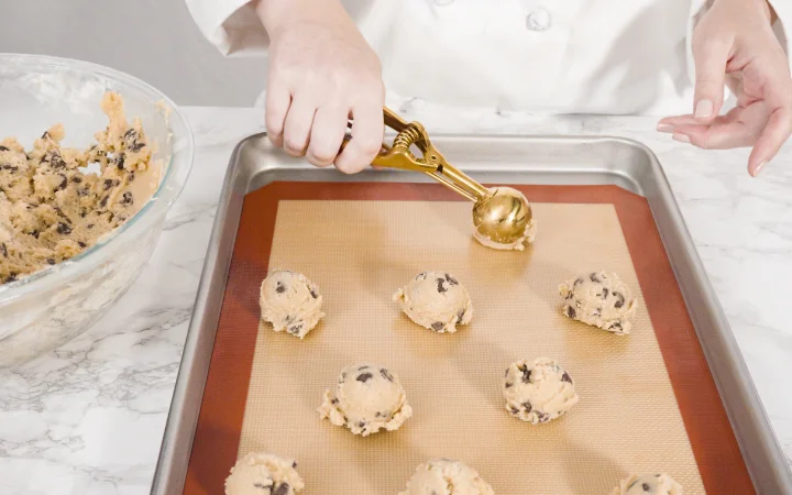 Close-up of hands skillfully mixing chocolate chip cookie dough in a bowl, highlighting the texture and consistency of the dough. Baking tools and a recipe card are visible in the background, emphasizing the art and science of dough preparation.