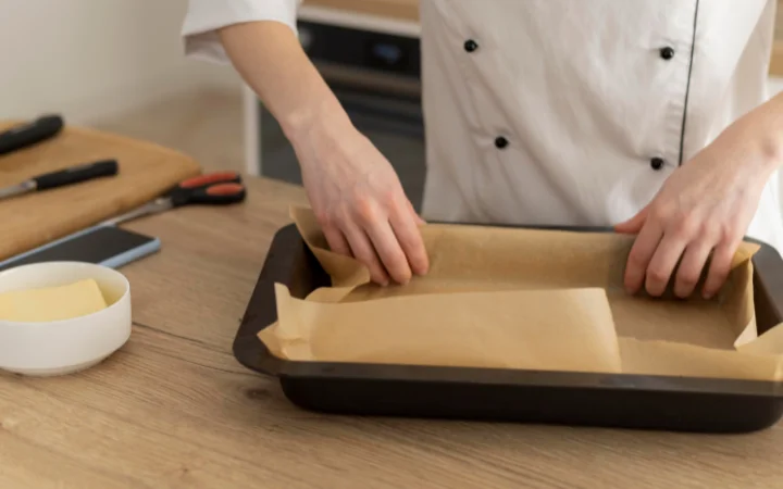 A home baker looking puzzled while examining a tray of flat chocolate chip cookies, with a kitchen background.