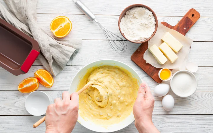 A baker's hands creaming butter and sugar in a mixing bowl, with a preheated oven in the background and a tray of Cowboy Cookie dough ready to bake.