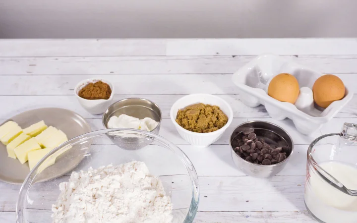  neatly arranged set of ingredients for Cowboy Cookies, including oats, pecans, coconut flakes, and chocolate chips, on a kitchen counter. Each ingredient is in its own bowl, showcasing their unique textures and colors.