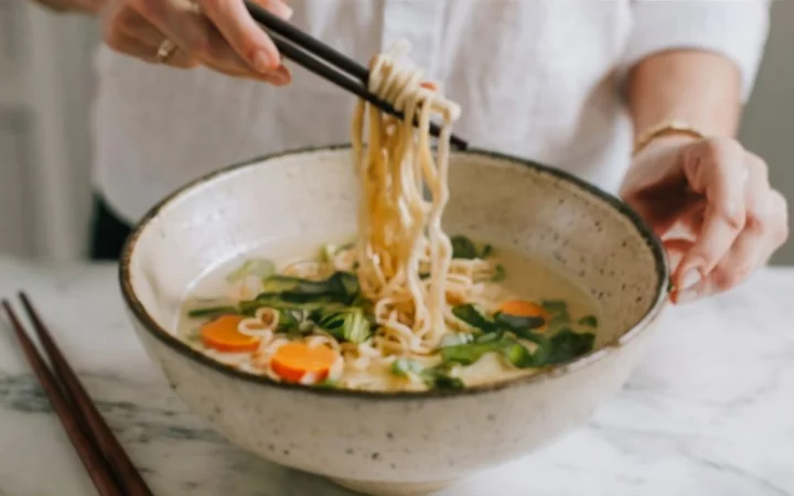 a step-by-step cooking process of ramen in a bright, airy kitchen, from boiling water in a pot to adding noodles and finally serving them in a beautiful ceramic bowl.