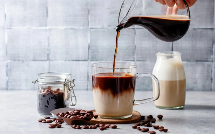  Ingredients for peppermint mocha laid out on a wooden kitchen counter, including fresh mint, a block of dark chocolate, a bag of espresso beans, milk, and sugar. A hand is seen pouring the melted chocolate into the milk.