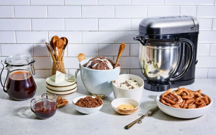 A rustic kitchen counter with ingredientst like: heavy cream, whole milk, granulated sugar, vanilla extract, toasted pecans, Turtle candies, caramel sauce, and hot fudge, alongside an ice cream maker.