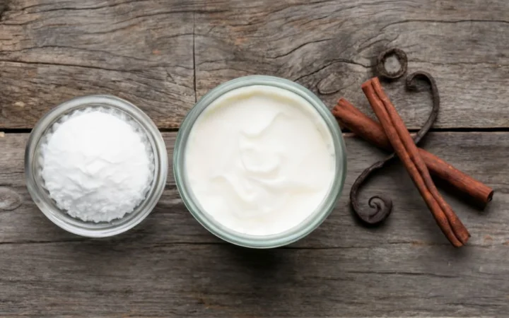 A flat lay of the three essential ingredients for making heavy whipping cream: a bottle of heavy cream, a bowl of confectioners' sugar, and a vanilla pod, arranged neatly on a rustic wooden surface