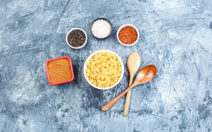 Top view ditalini pasta in white bowl with wooden spoons, spices on grey plaster background. horizontal