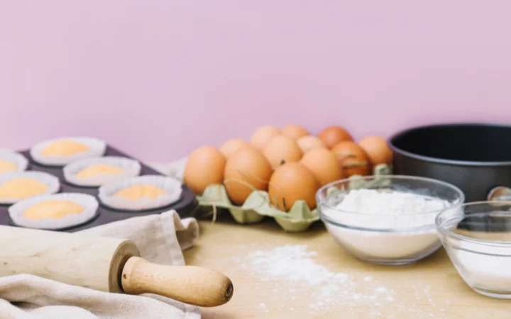 Baking cupcake ingredients with rolling pins on kitchen worktop against pink background