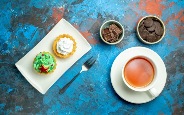 Top view a cup of tea cakes on plate chocolates in small bowls on blue red surface