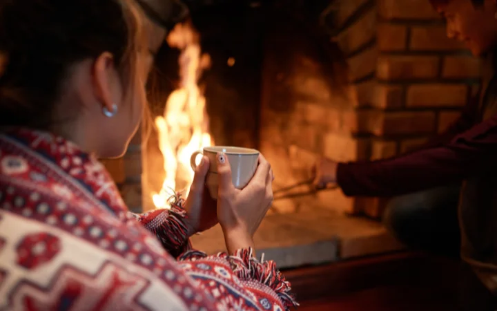 Over the shoulder shot of woman warming her hands on the mug sitting at the fireplace, her boyfriend dealing