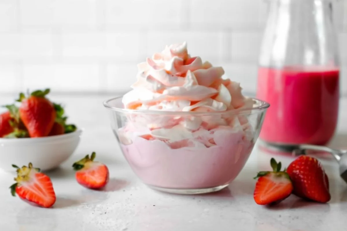 A light and airy bowl of strawberry whipped cream, with a vibrant pink hue, sitting on a kitchen counter beside a basket of fresh strawberries and a whisk.