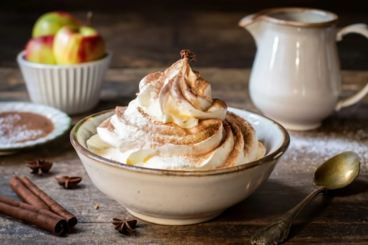 A light and airy bowl of whipped cream, with a sprinkle of espresso powder on top, sitting next to a decadent chocolate cake and a warm apple pie, all on a rustic kitchen table.